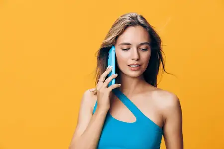 Стильная женщина в голубом топе разговаривает по смартфону на фоне ярко-желтой стены