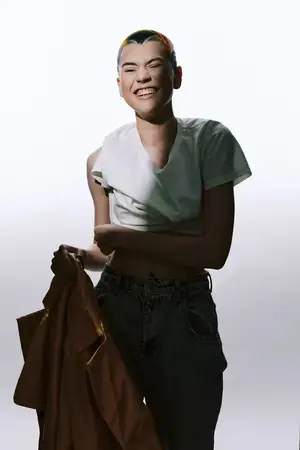 Беззаботная молодая женщина с короткими яркими волосами, радостно смеющаяся, одетая в повседневную белую рубашку и темные джинсы, держит коричневую куртку, изолирована на белом фоне, энергичное…