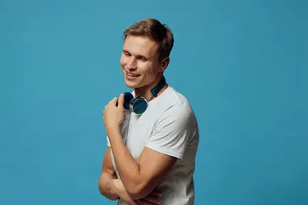 Улыбающийся молодой человек в белой футболке держит беспроводные наушники с закрытыми глазами на фоне синего фона, выражая расслабление и перерыв от гаджетов для психического здоровья
