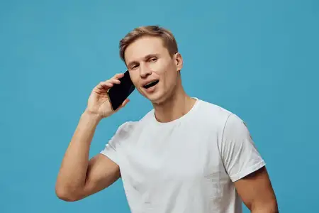 Молодой мужчина европеоидной расы разговаривает по смартфону, улыбаясь, счастливый и уверенный, одет в однотонную белую футболку, изолирован на синем фоне