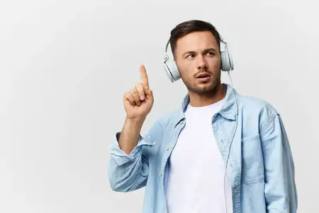 Задумчивый, задумчивый, взволнованный загорелый красивый мужчина в повседневной простой футболке и наушниках поднимает пальцы вверх, позируя изолированно на белом студийном фоне
