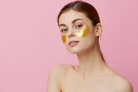 Молодая женщина с патчами для лица, ухаживающая за кожей, с обнажёнными плечами на розовом фоне