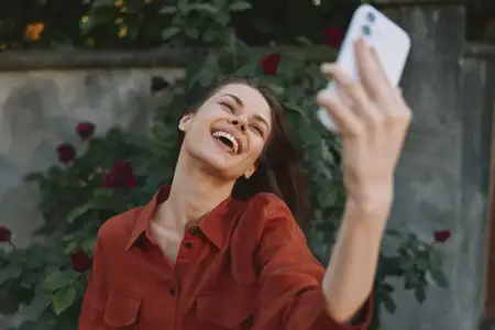Жизнерадостная женщина наслаждается солнечным днем на улице, запечатлевая спонтанный момент с помощью селфи на смартфон, одетая в повседневную одежду в садовой обстановке, с яркой и веселой атмосферой