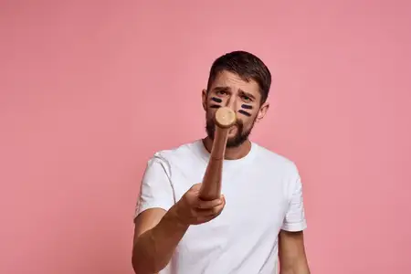 Эмоциональный мужчина с бейсбольной битой в руке на розовом фоне, на лице модели гримаса с чёрными линиями