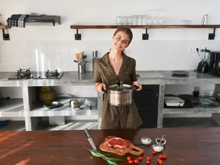 Женщина на кухне с кастрюлей в руках, с улыбкой готовит ужин из мяса и овощей на деревянном столе на фоне стильной кухни