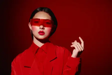Стильная молодая женщина в монохромном красном наряде и красных полупрозрачных очках уверенно позирует с яркими тёмными волосами на минималистичном фоне