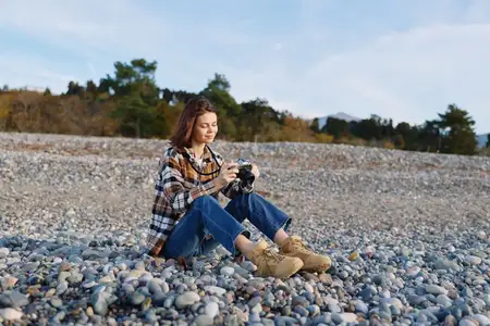 Женщина-фотограф в повседневной одежде на каменистом берегу, снимающая детали камерой
