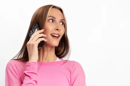 Улыбающаяся женщина в розовом свитере прижимает смартфон к уху и с радостным лицом смотрит в сторону