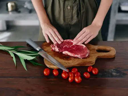 Женщина с ножом в руке нарезает свежее мясо для стейка для запекания на кухне, на столе соль, перец и другие специи, красные помидоры черри и зелень, приготовление ужина