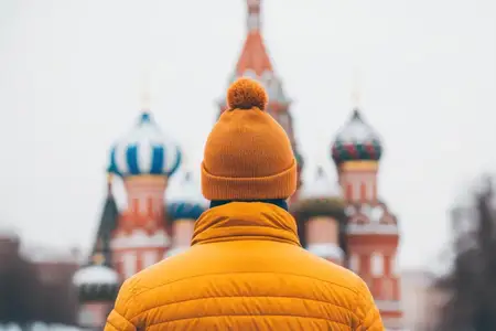 На этом изображении запечатлен молодой человек в ярком оранжевом зимнем пальто и шапке, смотрящий на знаменитый собор Василия Блаженного