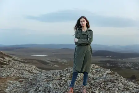 Уверенная в себе женщина в плаще стоит на вершине горы, скрестив руки на груди