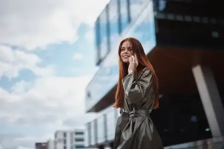 Молодая женщина-бизнесмен разговаривает по телефону с улыбкой, у неё рыжие волосы, она стоит на фоне высотных зданий в городе