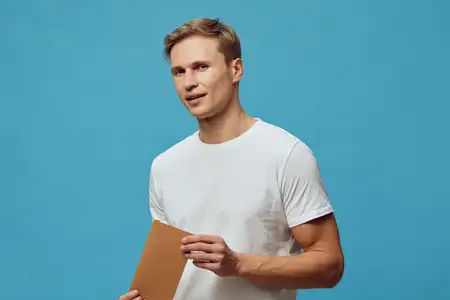 Расслабленный улыбающийся мужчина с аккуратной причёской в повседневной белой футболке держит коричневую папку или блокнот, уверенно позируя на однородном голубом фоне в студии