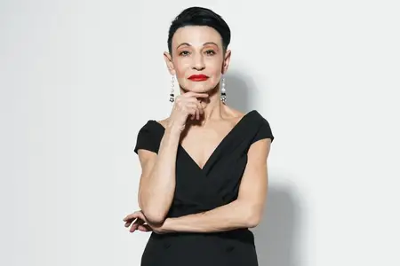 Красивая женщина в элегантном чёрном платье и с красной помадой позирует перед камерой на фоне белой стены