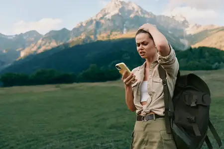 Лайфстайл-портрет женщины на природе с рюкзаком, использующей смартфон, пока она исследует горные пейзажи и зеленые луга, передающий независимость, спокойствие и активное благополучие на природе
