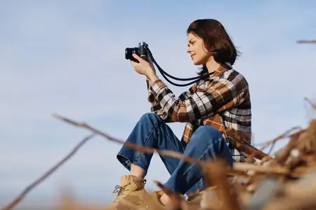 Женщина-фотограф в клетчатой рубашке и джинсах сидит среди ветвей на открытом воздухе, используя компактную камеру, чтобы запечатлеть безмятежную природную сцену и пейзаж с вниманием к деталям