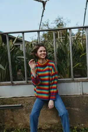 Улыбающаяся женщина в радужном полосатом свитере и синих джинсах сидит, прислонившись к перилам рядом с листвой теплицы