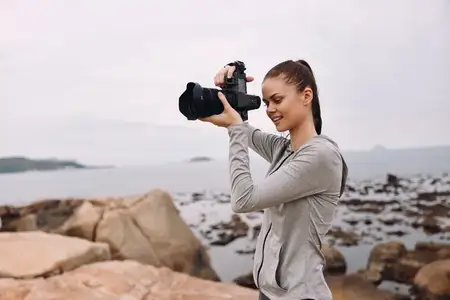Молодая женщина с радостным выражением лица фотографирует прекрасный океанский пейзаж на скалистом берегу, одетая в повседневную одежду, что отражает ее любовь к фотографии и природе