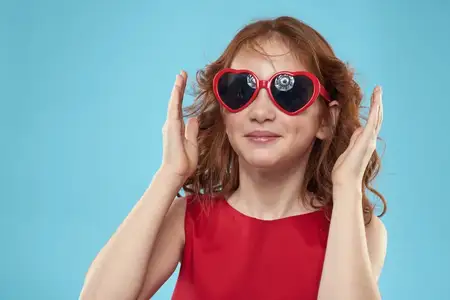 Маленькая девочка в солнечных очках в форме сердечек, красном платье, с вьющимися волосами на голубом фоне