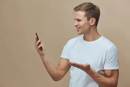 Счастливый молодой человек с короткими волосами держит смартфон и делает селфи, улыбаясь и жестикулируя свободной рукой, одетый в повседневную белую футболку