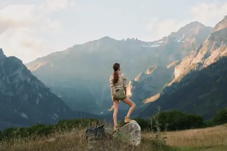 Молодая женщина с длинными волосами, в шортах и легкой рубашке, уверенно стоит на камне на травянистом горном лугу в окружении высоких вершин под частично облачным небом