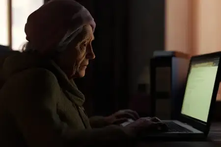 Пожилая женщина с седыми волосами и задумчивым выражением лица работает за ноутбуком дома, одетая в уютный вязаный свитер
