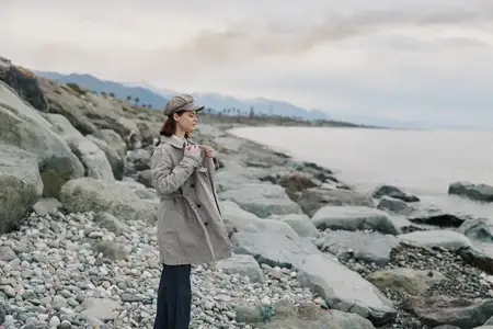 Женщина, одетая в клетчатое пальто и шляпу, стоит на каменистом пляже у спокойного моря