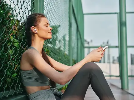 Молодая женщина в спортивной одежде наслаждается музыкой на телефоне, сидя у зелёного забора в безмятежной обстановке на открытом воздухе