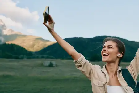 Улыбающаяся женщина в бежевой куртке делает селфи на смартфон на фоне живописного горного пейзажа