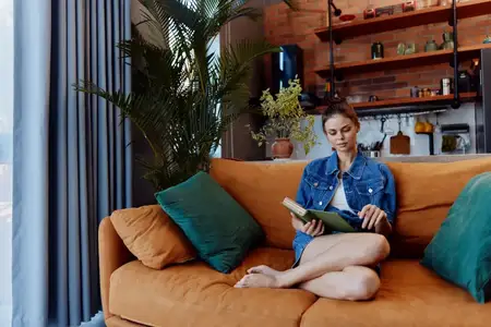 Женщина отдыхает на оранжевом диване у окна, читая книгу в уютной гостиной, куда проникает дневной свет