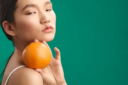 Азиатская женщина с гладкой кожей и темными волосами стоит с апельсином в руке, создавая минималистичное изображение, подчеркивающее цветовой контраст, баланс и естественную красоту