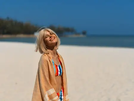 Счастливая женщина в полосатом купальнике, завернутая в полотенце, стоит на песчаном пляже под ясным голубым небом, выражая радость и расслабление в теплый летний день