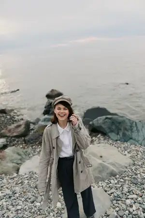 Улыбающаяся женщина в повседневном пальто и кепке позирует на галечном берегу на фоне океана