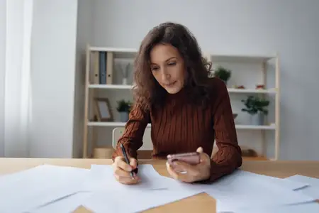 Серьёзная, несчастная, задумчивая кудрявая милая женщина сидит за столом с бумагами и документами, переживает из-за проблемы на работе, думает о неудаче, банкротстве, рассчитывает бюджет в телефоне