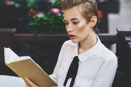 Эмоциональная женщина с короткими волосами читает книгу в ресторане, на заднем плане розовые цветы