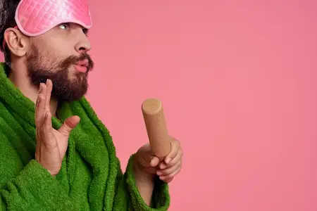 Мужчина в зелёном халате с розовой маской для сна на лице держит в руке скалку