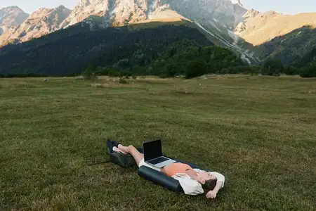 Одинокий работник отдыхает на широком лугу с ноутбуком, на фоне возвышающихся вдалеке гор и ясного голубого неба