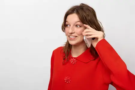Улыбающаяся молодая женщина в красном свитере держит смартфон у уха, глядя в сторону с жизнерадостным выражением лица