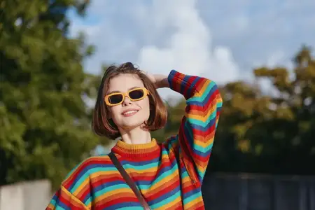 Женщина в радужном свитере и желтых солнцезащитных очках улыбается на улице, коротковолосая молодая взрослая откинулась назад в повседневной одежде с полосатым трикотажем, радостное настроение и…