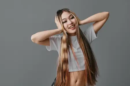 Девушка с длинными косичками позирует с закрытыми глазами и руками на голове в белом топе-кропе