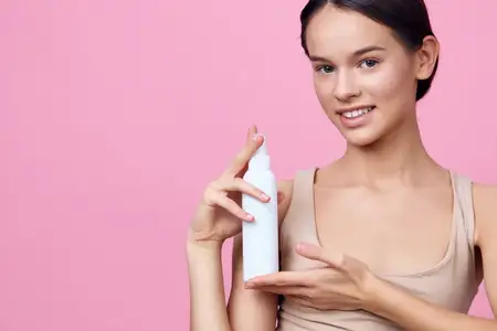 Улыбающаяся молодая женщина с естественным макияжем демонстрирует средство по уходу за кожей на ярко-розовом фоне, акцентируя внимание на красоте и здоровье, создавая свежее и юное настроение