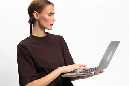 Молодая женщина в тёмной одежде работает на ноутбуке с серьёзным выражением лица на белом фоне
