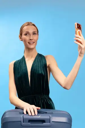 Женщина в стильном платье держит смартфон для селфи с чемоданом на фоне синего задника