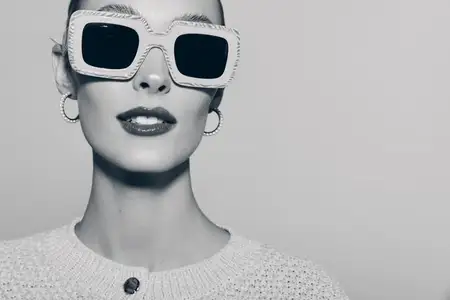 Модная белая женщина со светлой кожей и красными губами в больших квадратных солнцезащитных очках и серьгах-кольцах на монохромном студийном портрете