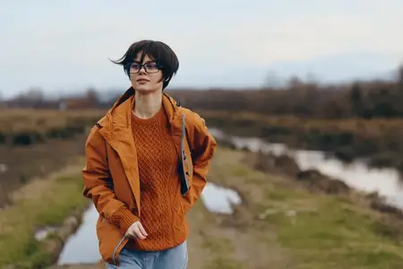 Женщина на открытом воздухе в стиле лайфстайл: речной пейзаж, куртка, свитер, очки, идет у реки, оранжевая куртка и вязаный свитер, очки, естественный дневной свет, мягкий фокус, луговой фон…