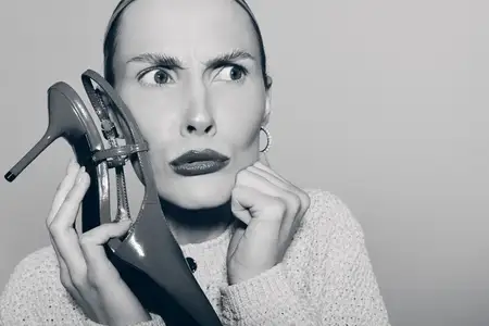 Бледнокожая женщина европеоидной расы в свитере с озадаченным выражением лица держит туфлю близко к лицу на нейтральном фоне