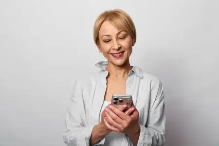 Женщина, смартфон, улыбка, общение, технологии, портрет, серый фон, повседневная одежда, пожилая, смотрит на экран мобильного телефона с легким смехом, расслабленная поза и дружелюбный фокус…