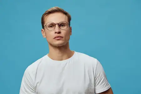 Серьезный молодой мужчина европеоидной расы в круглых очках, одетый в белую футболку, смотрит в камеру, стоя на голубом фоне
