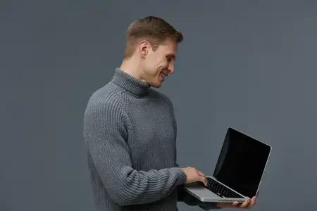Счастливый мужчина работает за ноутбуком, на нем уютный серый свитер, он стоит на фоне однотонного серого фона, наслаждается технологиями, повседневный стиль, настроение продуктивности или отдыха…