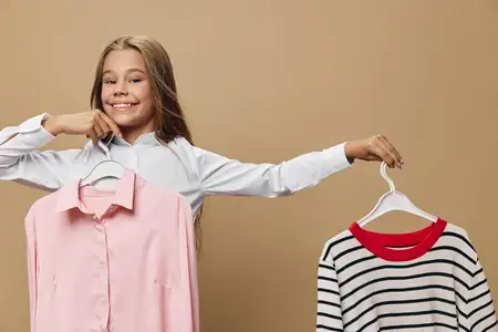 Улыбающаяся девочка-подросток держит одежду на вешалках: розовую рубашку и полосатый свитер